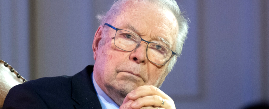 Krzysztof Zanussi w okularach, siedzi z dłońmi złożonymi, poważna mina.