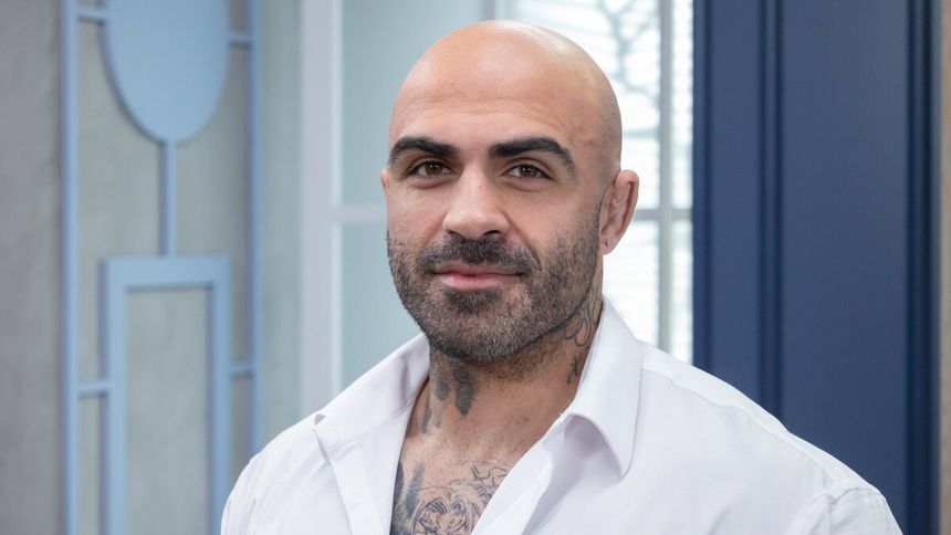 Akop Szostak z brodą i tatuażami, w białej koszuli, stoi w jasnym pomieszczeniu.