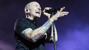 Mężczyzna z tatuażami, śpiewający do mikrofonu na scenie, z wyrazem radości na twarzy.