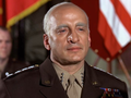 Mężczyzna w mundurze wojskowym, z odznaczeniami, na tle flag. Poważna ekspresja twarzy.