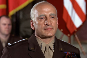 Mężczyzna w mundurze wojskowym, z odznaczeniami, na tle flag. Poważna ekspresja twarzy.