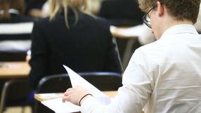 Uczestnik egzaminu siedzi przy biurku z kartkami w ręku, widoczne plecy innych osób.