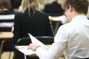 Uczestnik egzaminu siedzi przy biurku z kartkami w ręku, widoczne plecy innych osób.