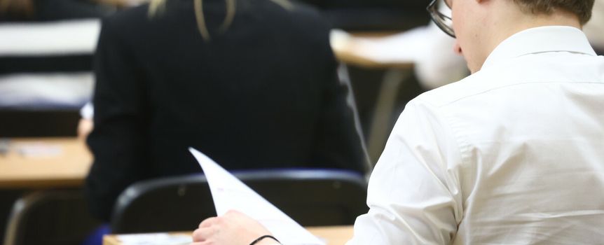 Uczestnik egzaminu siedzi przy biurku z kartkami w ręku, widoczne plecy innych osób.