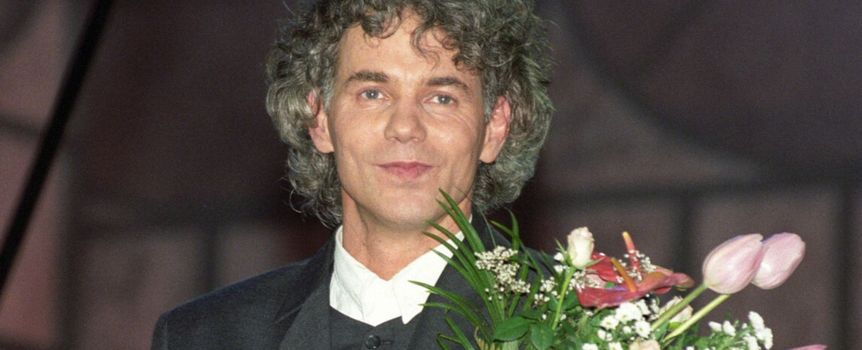 Mężczyzna z kręconymi włosami, w garniturze, trzyma bukiet kwiatów.