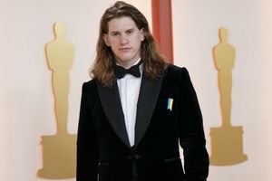 Mężczyzna w eleganckim czarnym garniturze z odznaką, stoi na tle figur oscarowych.