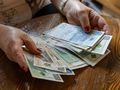 Ręka z dłońmi trzyma różne banknoty, część z nich leży na stole. Niewielka ilość monet obok.