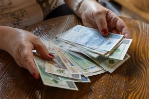 Ręka z dłońmi trzyma różne banknoty, część z nich leży na stole. Niewielka ilość monet obok.