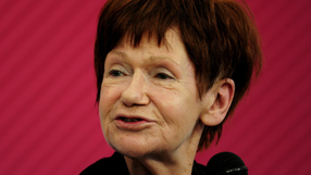 Zdjęcie Marii Czubaszek z krótkimi, rudymi włosami, uśmiechającej się do mikrofonu na różowym tle.