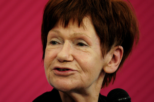 Zdjęcie Marii Czubaszek z krótkimi, rudymi włosami, uśmiechającej się do mikrofonu na różowym tle.