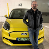 Mężczyzna w skórzanej kurtce stoi obok żółtego samochodu Ford w parkingu.