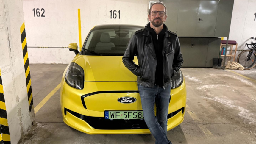 Mężczyzna w skórzanej kurtce stoi obok żółtego samochodu Ford w parkingu.