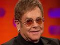 Elton John w okularach przeciwsłonecznych, uśmiechający się na tle kolorowej ścianki.