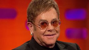 Elton John w okularach przeciwsłonecznych, uśmiechający się na tle kolorowej ścianki.
