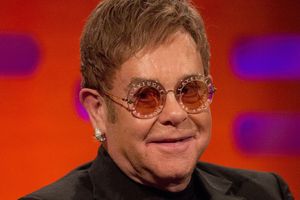Elton John w okularach przeciwsłonecznych, uśmiechający się na tle kolorowej ścianki.