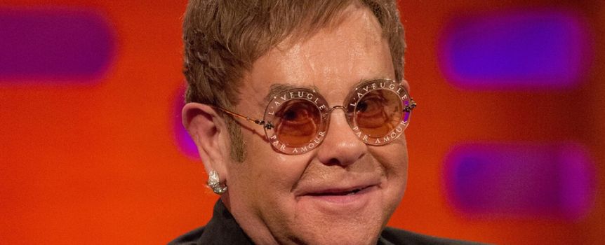 Elton John w okularach przeciwsłonecznych, uśmiechający się na tle kolorowej ścianki.