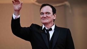 Mężczyzna w garniturze wita publiczność, uśmiechając się i unosząc rękę w geście pozdrowienia.