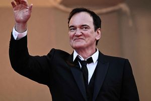 Mężczyzna w garniturze wita publiczność, uśmiechając się i unosząc rękę w geście pozdrowienia.