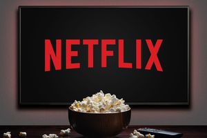 Zdjęcie przedstawia ekran z logo Netflix oraz miskę popcornu na stole.