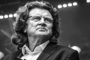 Zbigniew Wodecki. Mężczyzna z długimi włosami, w garniturze, patrzy w stronę widowni, na tle świecących świateł.