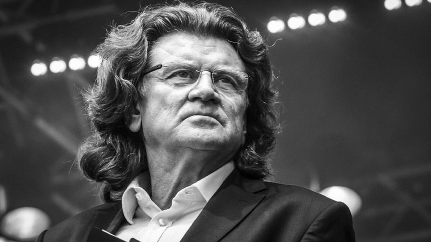 Zbigniew Wodecki. Mężczyzna z długimi włosami, w garniturze, patrzy w stronę widowni, na tle świecących świateł.