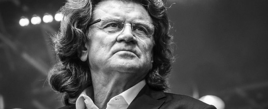 Zbigniew Wodecki. Mężczyzna z długimi włosami, w garniturze, patrzy w stronę widowni, na tle świecących świateł.