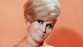 Kobieta z fryzurą w stylu lat 60., w pomarańczowej bluzce, na tle pomarańczowej ściany.