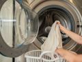 Osoba wkłada jasny ręcznik do pralki, obok biała koszykówka na pranie.