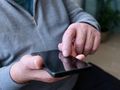 Mężczyzna w szarej bluzie dotyka ekranu smartfona jedną ręką, siedząc w pomieszczeniu.