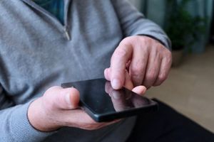 Mężczyzna w szarej bluzie dotyka ekranu smartfona jedną ręką, siedząc w pomieszczeniu.