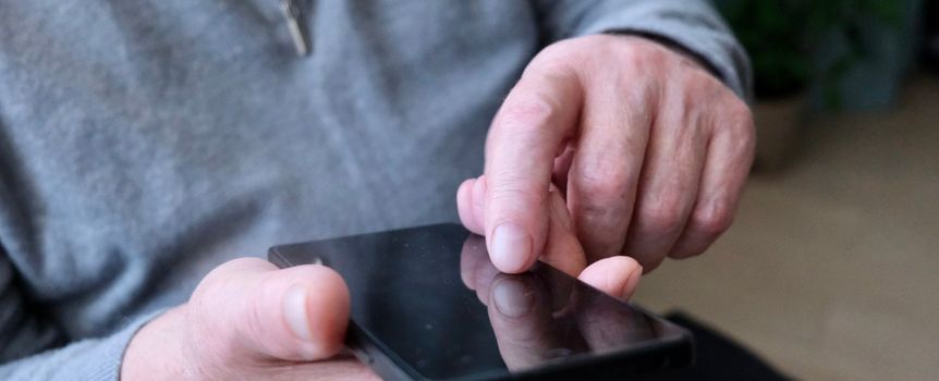 Mężczyzna w szarej bluzie dotyka ekranu smartfona jedną ręką, siedząc w pomieszczeniu.