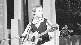 Andrzej Rosiewicz z gitarą na scenie, ubrany w kamizelkę i biały krawat. Czarno-białe zdjęcie.