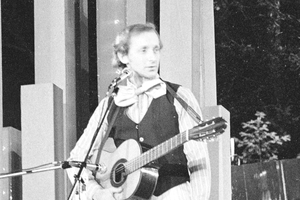 Andrzej Rosiewicz z gitarą na scenie, ubrany w kamizelkę i biały krawat. Czarno-białe zdjęcie.