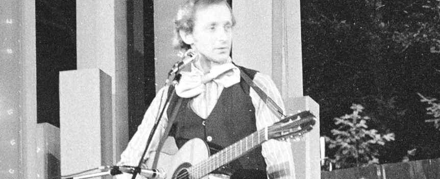 Andrzej Rosiewicz z gitarą na scenie, ubrany w kamizelkę i biały krawat. Czarno-białe zdjęcie.