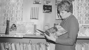 Kobieta w swetrze przegląda książkę w bibliotece, w tle regał z literaturą.