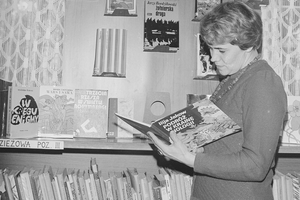 Kobieta w swetrze przegląda książkę w bibliotece, w tle regał z literaturą.