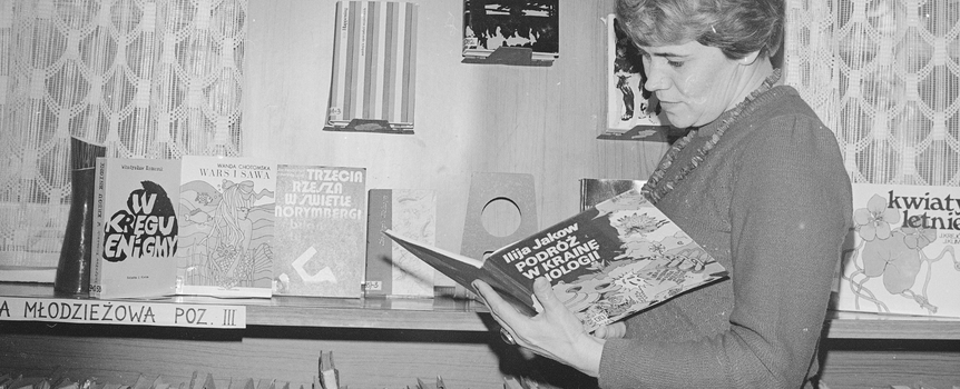 Kobieta w swetrze przegląda książkę w bibliotece, w tle regał z literaturą.