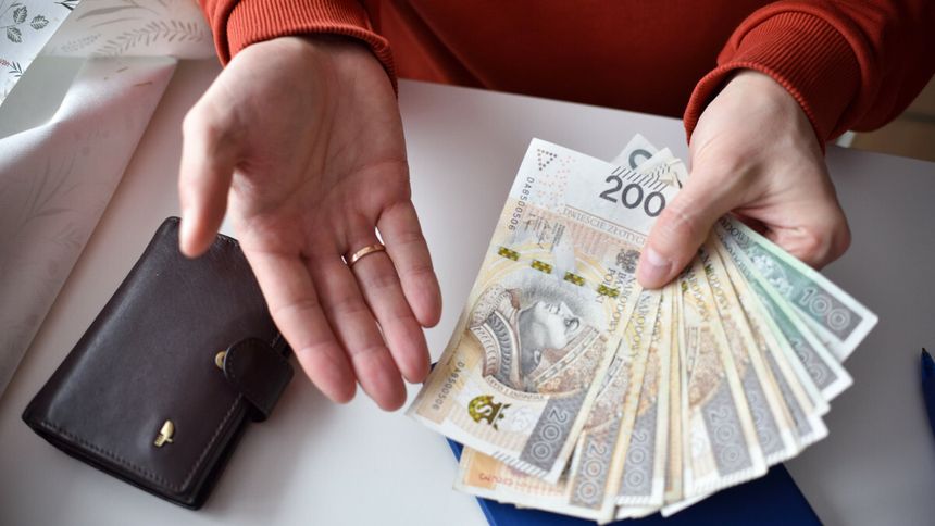Osoba trzymająca banknoty w dłoni, obok portfel oraz kartki na stole. Na banknotach są różne nominały.