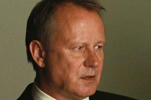 Mężczyzna w ciemnym garniturze z poważnym wyrazem twarzy, w tle niewyraźne kolory.