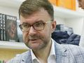 Jacek Dukaj. Mężczyzna w okularach, w jasnej marynarce, rozmawia z osobą z lewej strony, w tle książki.