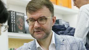 Jacek Dukaj. Mężczyzna w okularach, w jasnej marynarce, rozmawia z osobą z lewej strony, w tle książki.