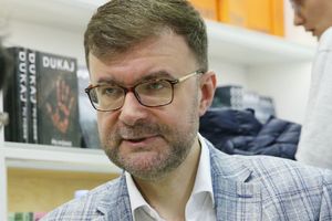 Jacek Dukaj. Mężczyzna w okularach, w jasnej marynarce, rozmawia z osobą z lewej strony, w tle książki.