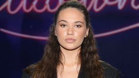 Na zdjęciu młoda kobieta z długimi, brązowymi włosami, patrzy w kamerę. Tło w ciemnych kolorach.