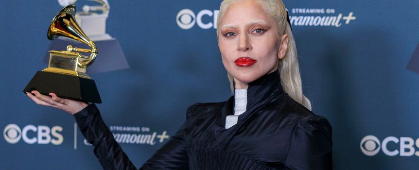 Lady Gaga. Artystka z białymi włosami, trzymająca statuetkę Grammy na tle niebieskiego tła.