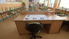 Pusty nauczycielski stół w klasie, dookoła odwrócone krzesła, jasne ściany.