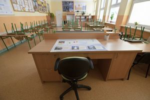 Pusty nauczycielski stół w klasie, dookoła odwrócone krzesła, jasne ściany.