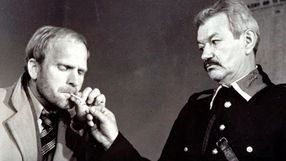 Dwóch mężczyzn w starych ubraniach, jeden zapala papierosa drugiemu. Czarno-biała scena z dramatem.