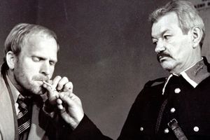Dwóch mężczyzn w starych ubraniach, jeden zapala papierosa drugiemu. Czarno-biała scena z dramatem.