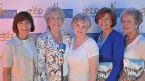 Na zdjęciu pięć uśmiechniętych kobiet, pozujących przed kolorowym tłem. Trzymają albumy.