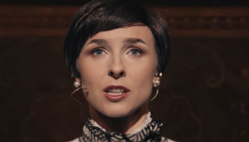 Kobieta z krótkimi włosami, w eleganckiej bluzce, mówi do publiczności na scenie.